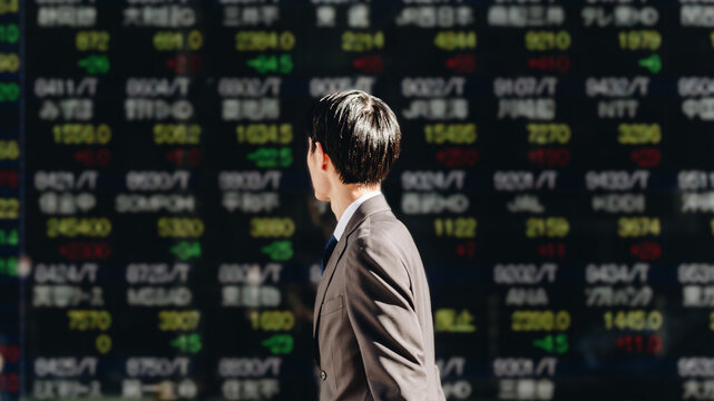 NISAでの株の運用に興味のある若いビジネスマンの男性が屋外の電光掲示板の株価ボードを見るイメージ