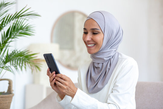 A beautiful woman in a hijab is using the phone close up, a Muslim woman is sitting on the sofa in the living room, holding a smartphone in her hands, browsing the Internet and smiling joyfully.