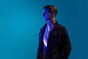 Portrait of handsome young man in stylish clothes, posing with serious expression over blue background in neon light. Digital light reflection on body. Concept of modern art, cyberpunk, creativity