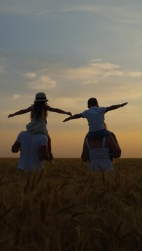 Vertical Screen: Slow motion mom and dad carrying kids on shoulders while walking in wheat field at sunset, boy and girl spreading arms as if flying, blue sky on background. Happy family together