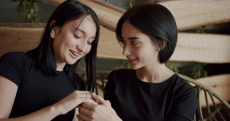 Couple, lgbt and lesbian women at home. Low angle handheld shot of young lesbian couple smiling and touching hands gently while sitting on sofa against window in light living room at home.