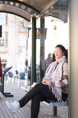 Young Asian girl with glasses listening to music through headphones while waiting for the bus. Concept of transportation and mobility.