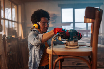 Female carpenter sanding chair with power tools.
