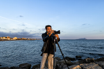Fototapeta premium Photographer and travel director on a seaside shooting with models looks into the viewfinder to take the best photo at sunset. Conceptual photo of study on photography