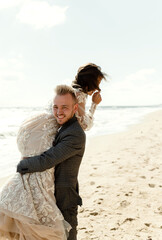 bearded groom with a smile carries the bride. newlyweds are having fun on the beach. wedding couple on the sea cute hugging