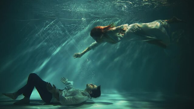 Fantasy gothic romantic couple fall in love man king and woman fashion model posing under water dark blue sea. Wet white dress. Fairy tale sexy girl river nymph swims saves drowning guy. Video shoot