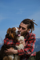 Young Caucasian man with dreadlocks sits on hilltop with dog and hugs and kisses. Australian Shepherd on hike with male owner. Close up portrait. Concept of mountain tourism and travel.