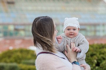 A young mother holds a charming little son in her arms. The kid looks at the camera and smiles. Portrait of a beautiful chubby 4-month-old baby.