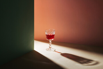 Creative scene crystal glass with red wine and shadow. Minimal party concept on beige, pink and...