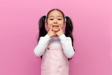 little Asian girl in pink sundress with long hair points her fingers at smile and healthy teeth, Korean preschool child