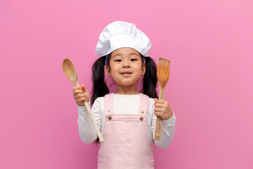 little asian girl in chef uniform holding kitchen items on pink isolated background, korean child...