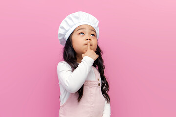 little pensive asian girl in chef uniform pointing finger up and thinking over question on pink...