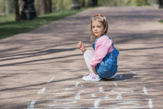 A happy girl in pink clothes draws classics with chalk on the asphalt on the street. Portrait of a little girl playing and jumping hopscotch on a sunny summer day. Creative development of children.