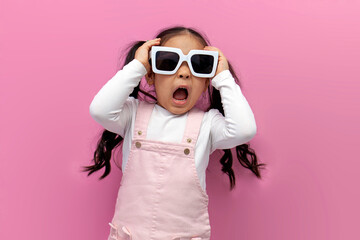 little shocked preschool girl in white sunglasses and pink sundress screams and is surprised on pink background