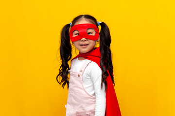 little asian girl in superman costume and mask smiles, korean child in superhero cape on yellow...