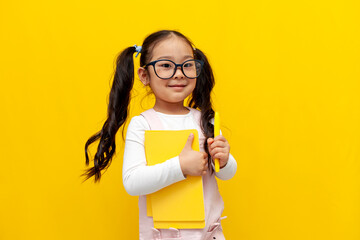 little asian girl with glasses holding notebooks and pen and smiling on yellow isolated background