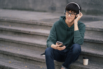 Attractive happy young Asian student sitting on the steps of the university, using smartphone and headphones having a video chat.