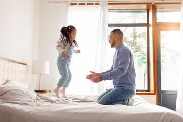 little asian girl jumping on bed at home with dad, korean man playing with daughter, happy family,...