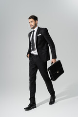 full length of businessman in black pantsuit walking with briefcase on grey background. - obrazy, fototapety, plakaty