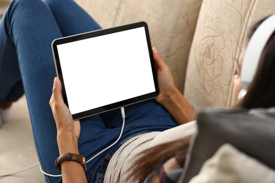 Mockup image of a digital tablet with black screen in hands of woman in headphones. Person resting at home on sofa with tablet over shoulder view