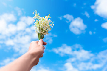 A woman's hand holds a bouquet of lilies of the valley against a blue sky