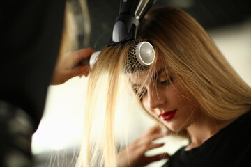 Drying blond hair with a hairdryer and a round brush in barbershop. Master doing hair styling to a client in beauty salon