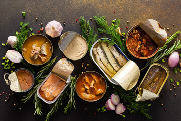 Different open tin cans with canned fish among spices and herbs on a brown background, canned...