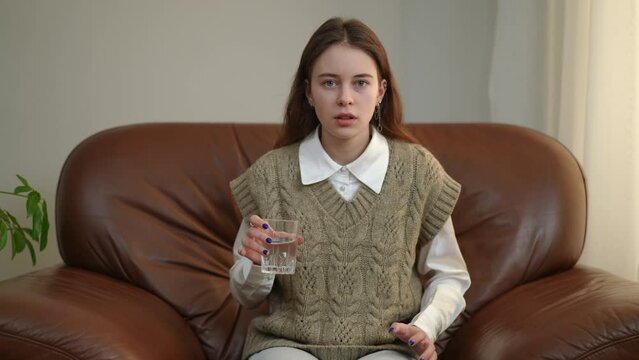 Front view scared nervous teenage girl looking at camera with glass of water shaking in hand. Medium shot portrait of frightened stressed Caucasian teenager posing indoors