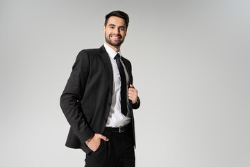 Obraz na płótnie Canvas bearded businessman in black suit standing with hand in pocket and smiling at camera isolated on grey.