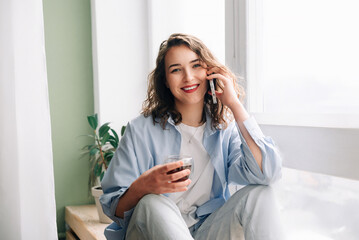 Happy woman chatting on phone with sister or friend over tea on kitchen windowsill. Pleasant conversation with loved ones. Joyful young woman having fun talking on phone while drinking tea at home