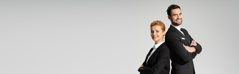 happy managers in black blazers smiling at camera while posing with folded arms isolated on grey, banner.