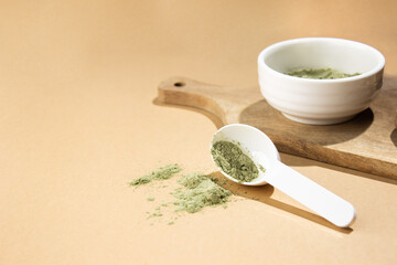 Green powder in a small white ceramic bowl, with a white spoon on a wooden board, on a beige...