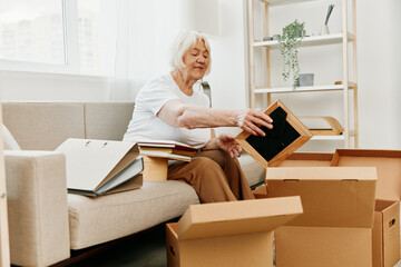 elderly woman sits on a sofa at home with boxes. collecting things with memories and moving and...