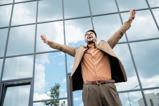 low angle view of successful overjoyed businessman in trendy clothes showing triumph gesture near modern city building.