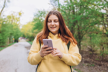 A portrait of a smiling beautiful woman walking with phone on nature background. Happy woman with backpack is using a smartphone in park outdoors, spring time. Traveler