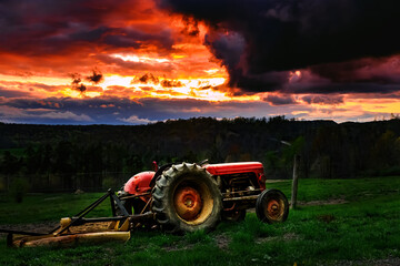 A red farm tractor is parked in the grass along the field with an amazing sunset behind it.  Strom...