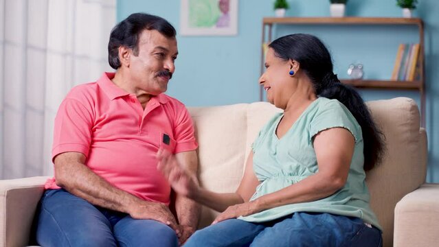 happy Indian senior couple discussing by talking each other while sitting on sofa at home - concept of communication, long relationship bonding and understanding.