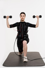 Fit girl with dumbbells in EMS suit that uses electrical impulses to stimulate muscles on white...