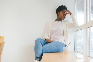 Sad woman in a cafe holding head with hand wearing formal jacket sitting on cafe windowsill. Negative person. Young woman in cozy cafe