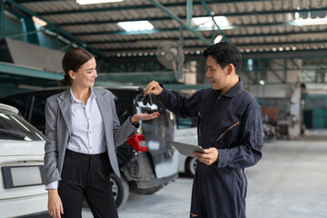 Businesswoman receiving car keys from mechanic man in workshop garage. Asian engineer man in uniform giving key to female client after checking and maintaining car engine at automotive service center.