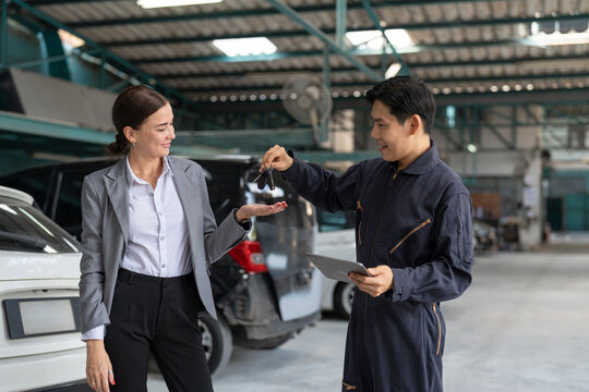 Businesswoman receiving car keys from mechanic man in workshop garage. Asian engineer man in uniform giving key to female client after checking and maintaining car engine at automotive service center.