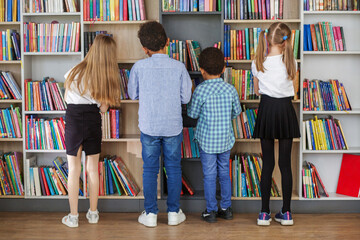 Four multietnic children look for books near bookshelves and read together in school library. Back