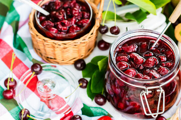 Cooked homemade cherry jam in glass jar on white wooden table outdoors, fresh cherry jam close up, food and healthy eating concept