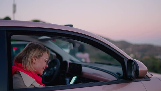 Young smiling millennial female photographing sunset view with mobile phone camera, sitting in rental car enjoying stop in travel. Young woman blogger sending text message in chat sharing impression