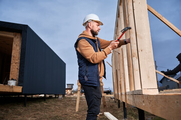 Man worker building wooden frame house on pile foundation. Carpenter hammering nail into wooden joist, using hammer. Carpentry concept.