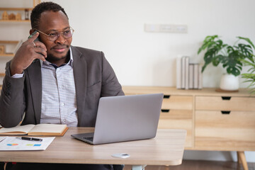 Obraz premium An African American businessman in a black suit uses a mobile phone to communicate with clients and a laptop to view company profits. to build confidence for customers to come and invest in the office