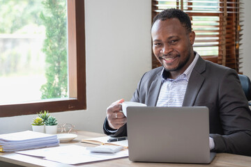 African American businessman in a black suit holding a cup of coffee to relax after checking company work on a laptop inside the office.