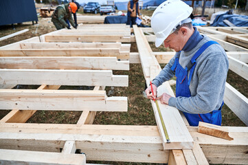 Fototapeta premium Man worker building wooden frame house on pile foundation. Carpenter using tape measure for measuring wooden planks and making marks with pencil. Carpentry concept.