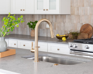 A kitchen faucet detail with a grey stone countertop island, gold faucet, stainless steel...