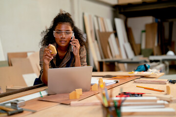 Carpenter woman use mobile phone to contact customer and discuss about wood product in front of laptop in workplace.